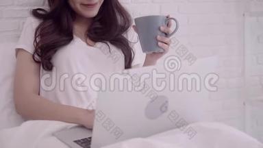 一幅美丽迷人的亚洲女人在床上躺着时，用电脑或笔记本电脑拿着一杯温暖的咖啡或茶的肖像。